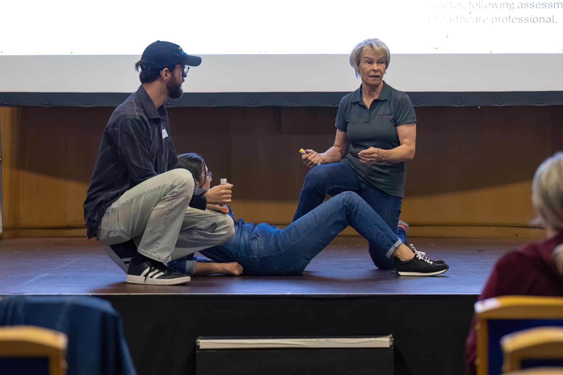 MMFTutorConference2026.23.web Three adults, identified as New TEMPO Ambassadors, demonstrate a first aid technique onstage; one person lies on their back, another kneels nearby explaining and holding a marker pen, while the third crouches, supporting the person on the floor. A projected screen is visible behind them.