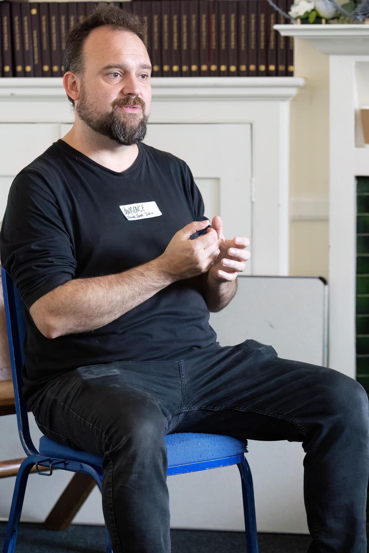 MMFTutorConference2026.25.web A man with a beard sits on a blue chair, wearing a black shirt and holding a marker, gesturing with his hands as he discusses the role of the New TEMPO Ambassadors. Shelves of books and a fireplace are visible in the background.