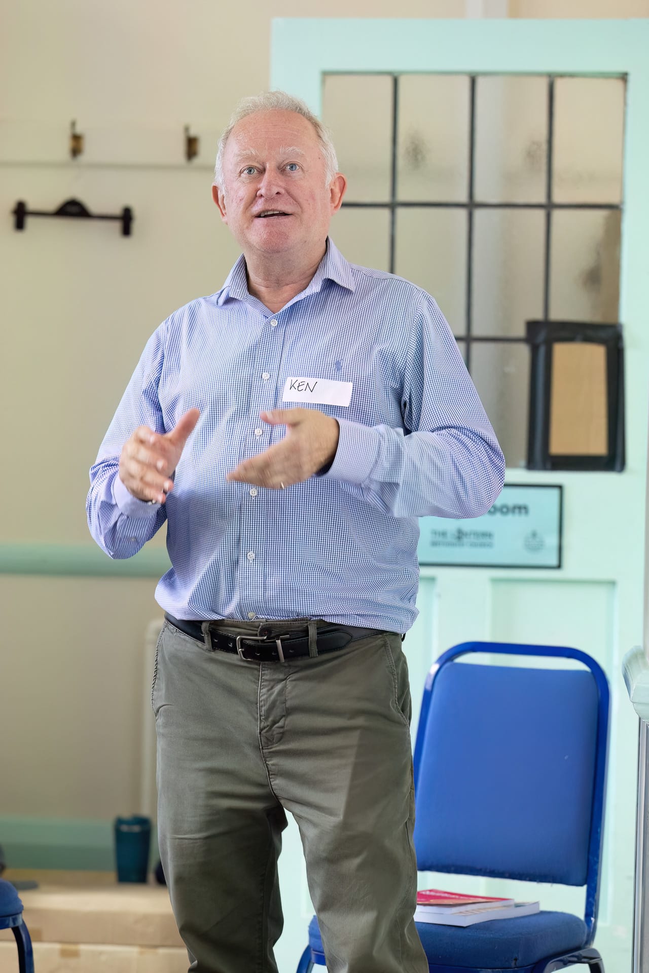 MMFTutorConference2026.29.web An older man in a blue shirt stands and gestures whilst speaking in a room with blue chairs and a pale blue door. He wears a name badge that says Ken and appears engaged in conversation or presentation, possibly addressing the group as one of the New TEMPO Ambassadors.