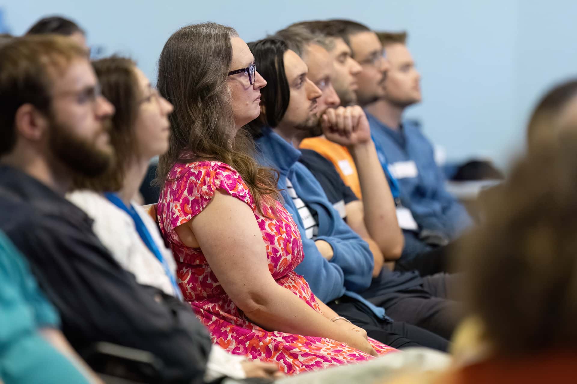 MMFTutorConference2026.37.web A group of people sits in rows, attentively listening to a speaker. At the centre, a woman in a bright pink dress with glasses is in focus—one of the New TEMPO Ambassadors. The background is softly blurred, highlighting the audience’s concentration.