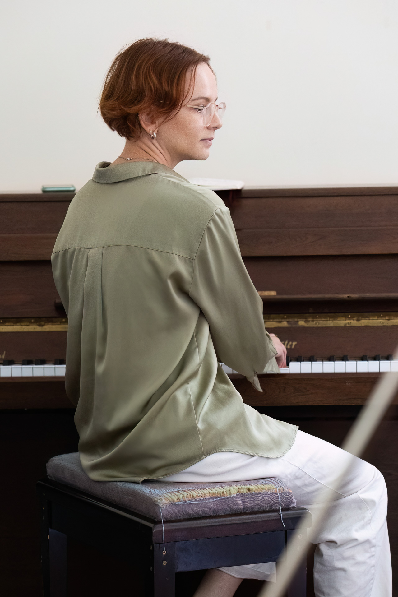 MMFTutorConference2026.44.web A person with short hair and glasses, wearing a light green shirt and white trousers, sits on a piano stool, turning to look over their shoulder while seated at an upright piano—one of the New TEMPO Ambassadors ready to inspire with their music.