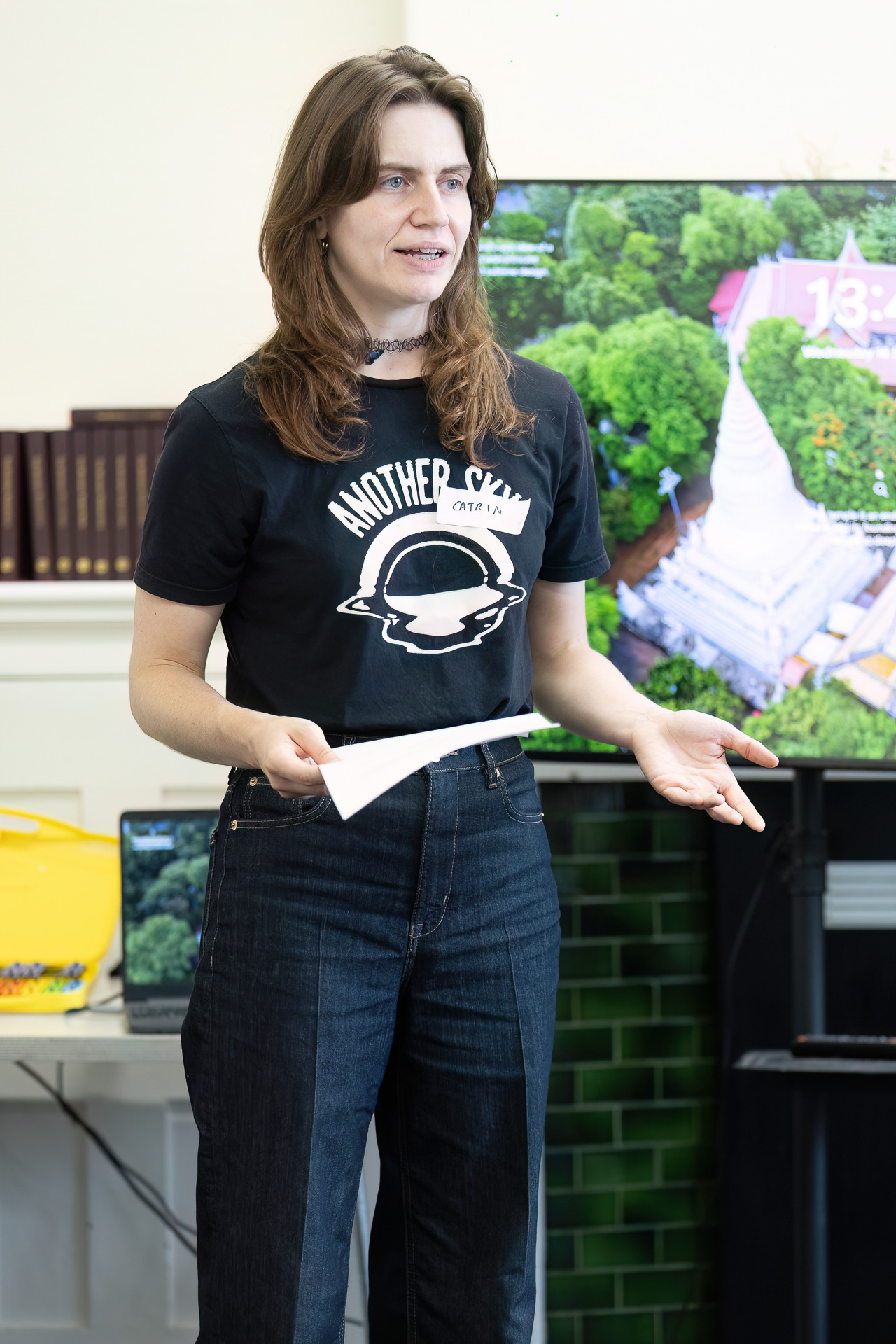 MMFTutorConference2026.46.web A person with long brown hair, wearing a black Another Sky T-shirt and jeans, speaks whilst holding papers in front of a TV displaying an aerial view of a white temple, introducing the new TEMPO Ambassadors.