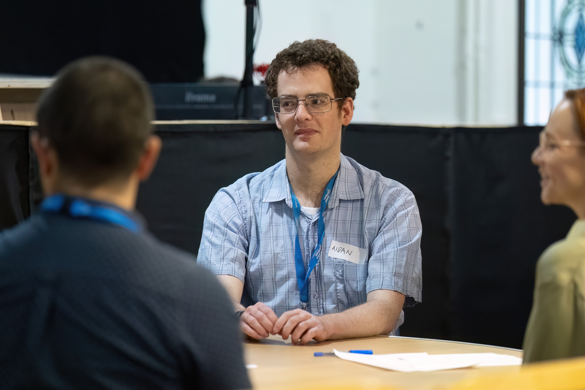 MMFTutorConference2026.57.web A man with short curly hair and glasses sits at a table during a meeting, wearing a name badge and short-sleeved checked shirt. Two other people, one male and one female, are partially visible facing him, as the group discusses their roles as New TEMPO Ambassadors.