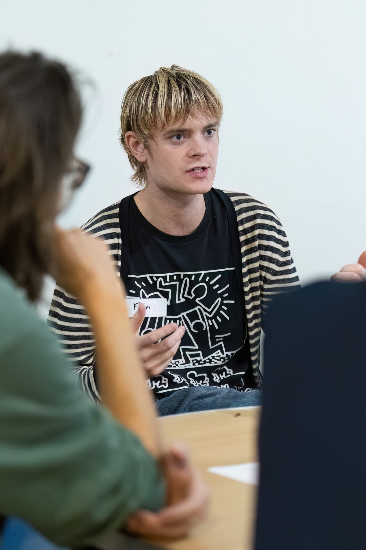 MMFTutorConference2026.60.web A young person with blond hair and a striped jumper gestures while speaking in a group discussion, sitting at a table with others—one of the New TEMPO Ambassadors actively engaging in the conversation.