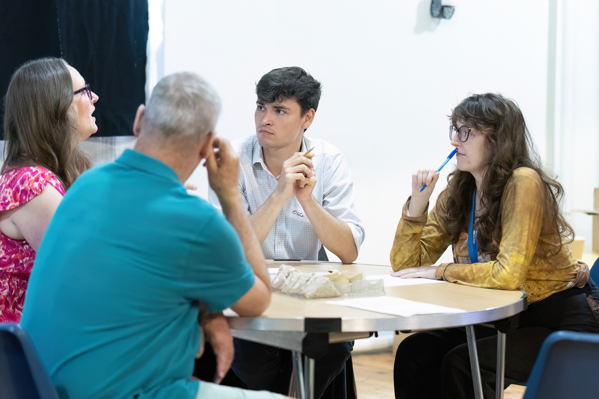 MMFTutorConference2026.62.web Four adults, including the New TEMPO Ambassadors, sit around a table engaged in discussion. Papers and models are on the table. One woman gestures whilst speaking, and the others listen intently, appearing thoughtful and focused.