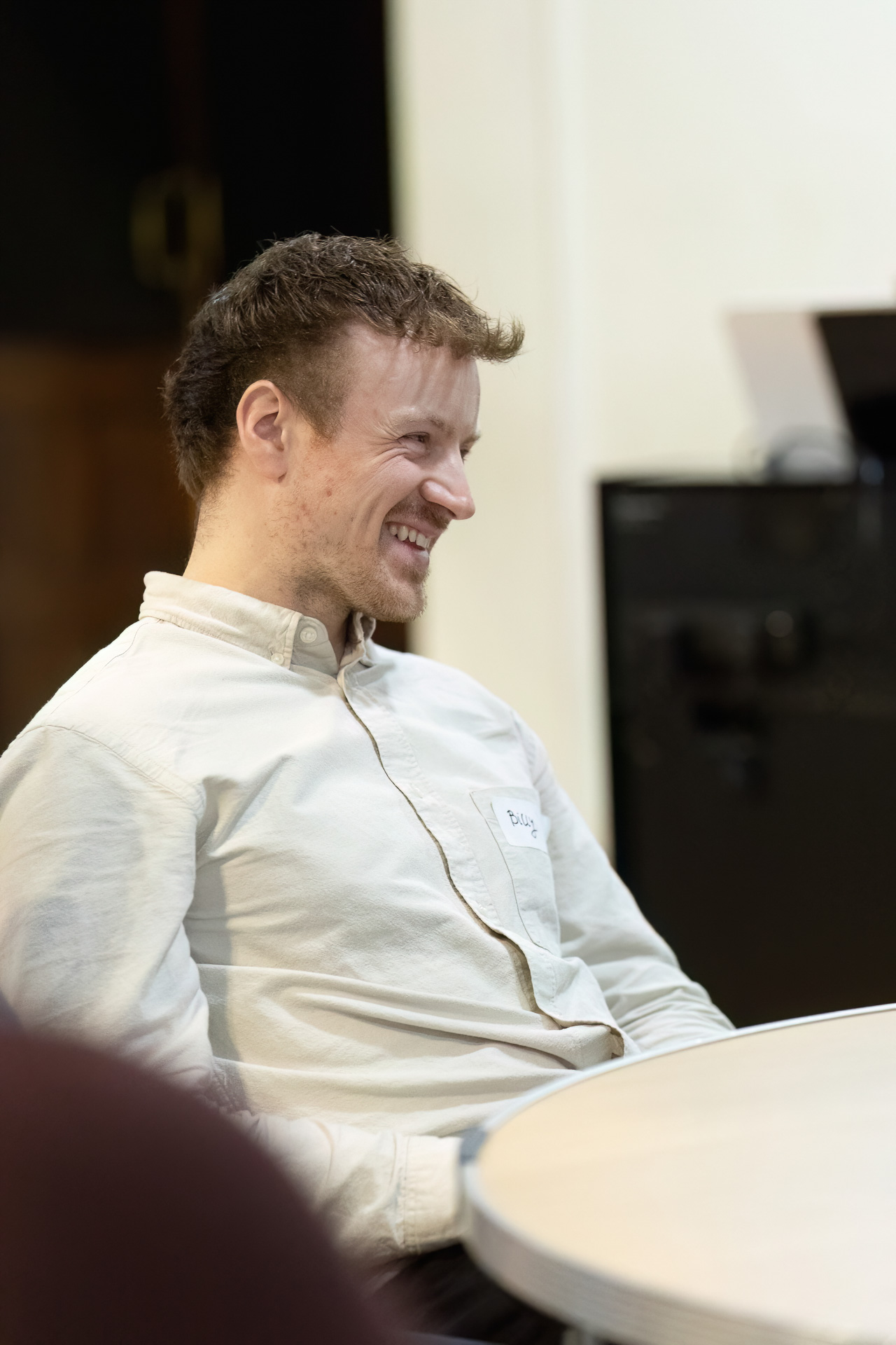 MMFTutorConference2026.65.web A man with short brown hair, wearing a white button-up shirt, sits at a round table and smiles, looking off to the side. A name tag is visible on his shirt, highlighting his role among the new TEMPO Ambassadors. The background is blurred.