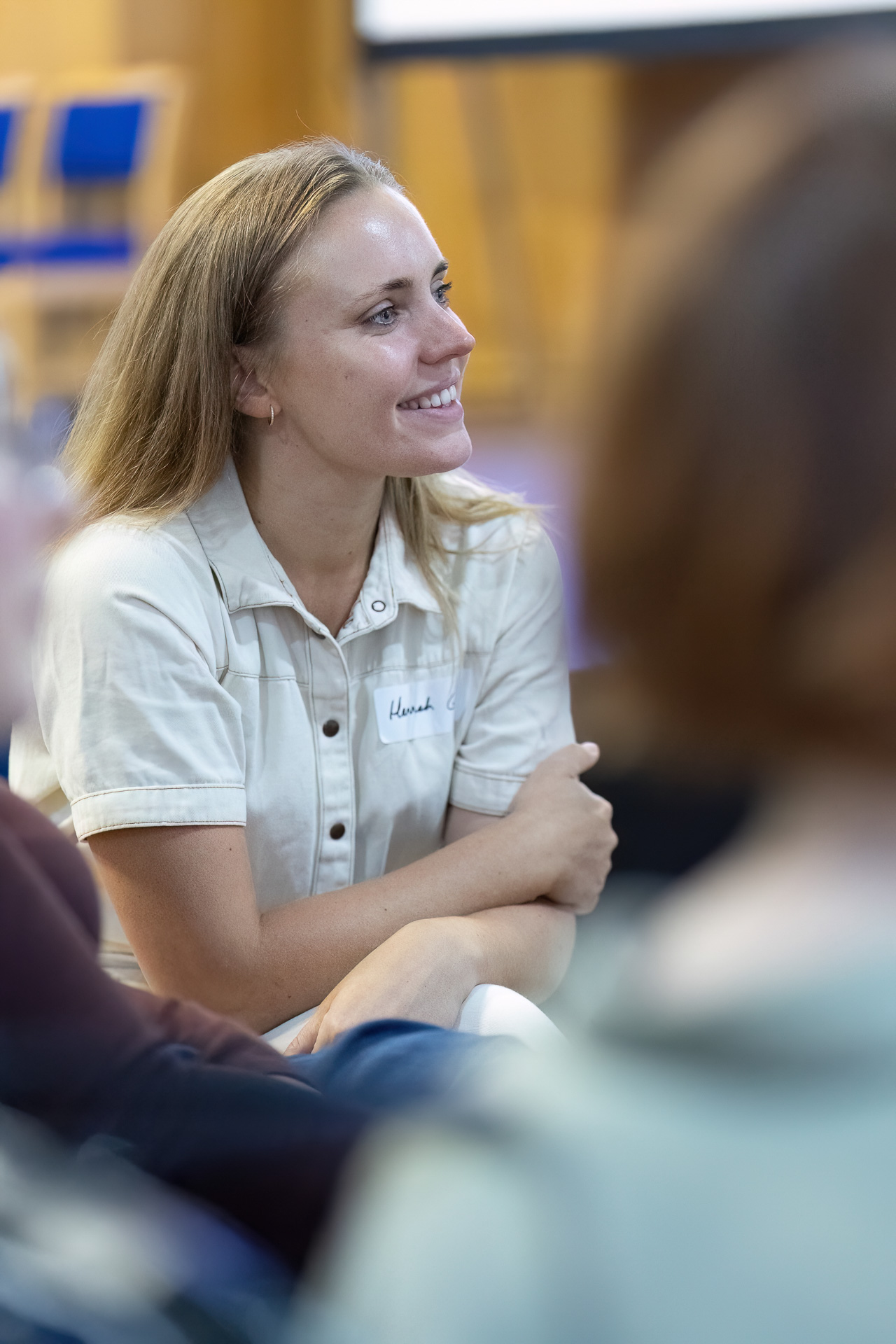 MMFTutorConference2026.68.web A woman with light brown hair, wearing a short-sleeved cream shirt and a name badge, sits smiling with her arms crossed, appearing engaged in an indoor event for the New TEMPO Ambassadors. Other people are blurred in the foreground.
