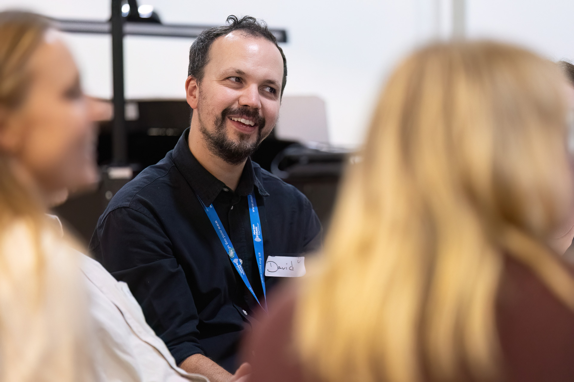 MMFTutorConference2026.74.web A man with a name badge sits and smiles while talking with two people in a brightly lit indoor setting, embodying the welcoming spirit of the new TEMPO Ambassadors.