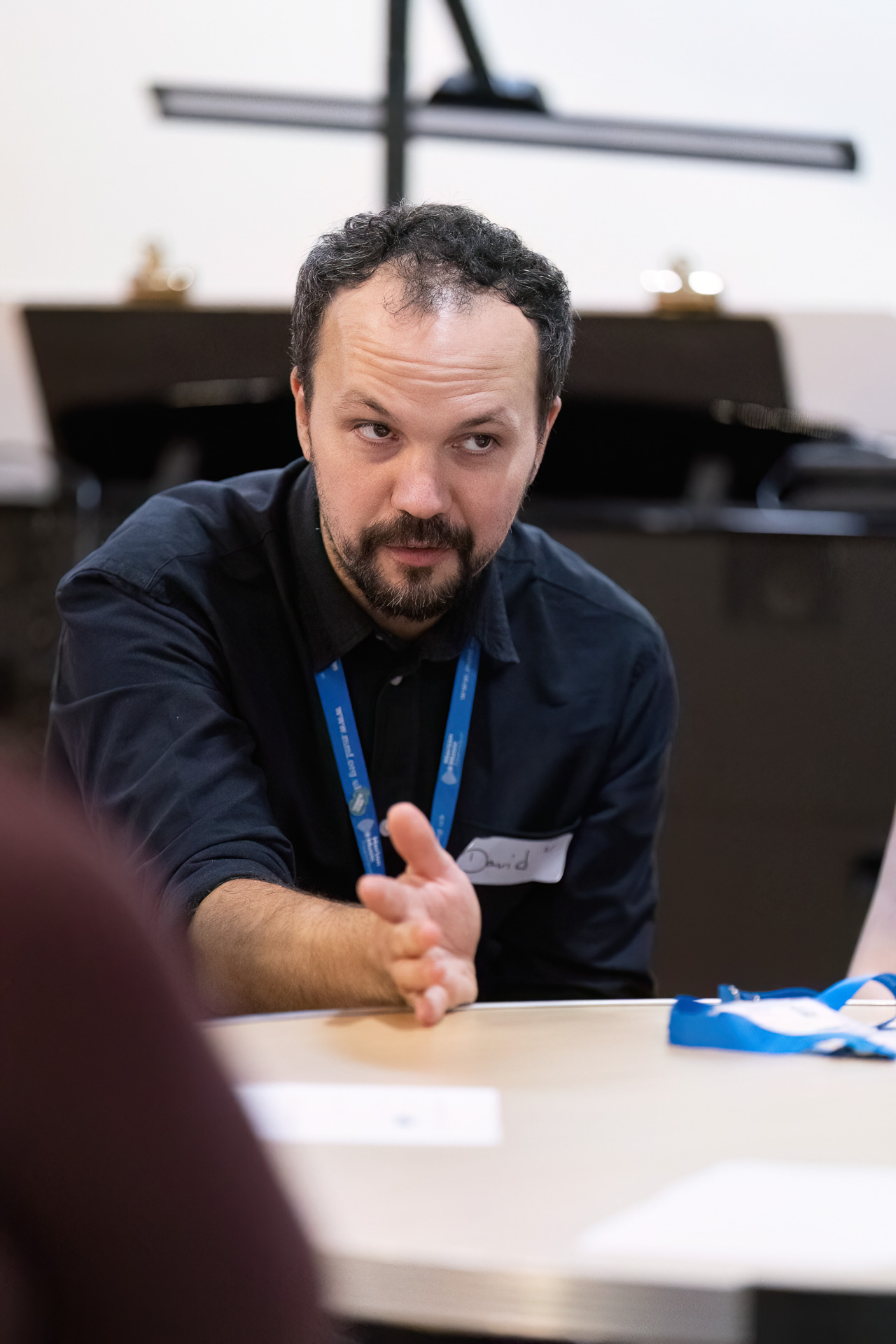 MMFTutorConference2026.76.web A man with a beard, wearing a dark shirt and blue lanyard, gestures with his hand whilst seated at a table, engaging in conversation as one of the new TEMPO Ambassadors.