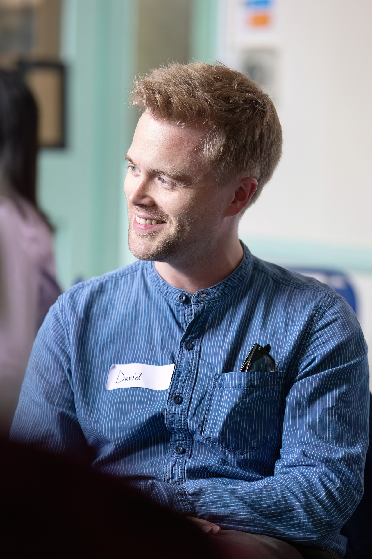 MMFTutorConference2026.81.web A man with fair hair and a beard, wearing a blue shirt with a name badge that says David, smiles while sitting indoors; a pair of spectacles is in his shirt pocket. He looks ready to represent the New TEMPO Ambassadors programme.