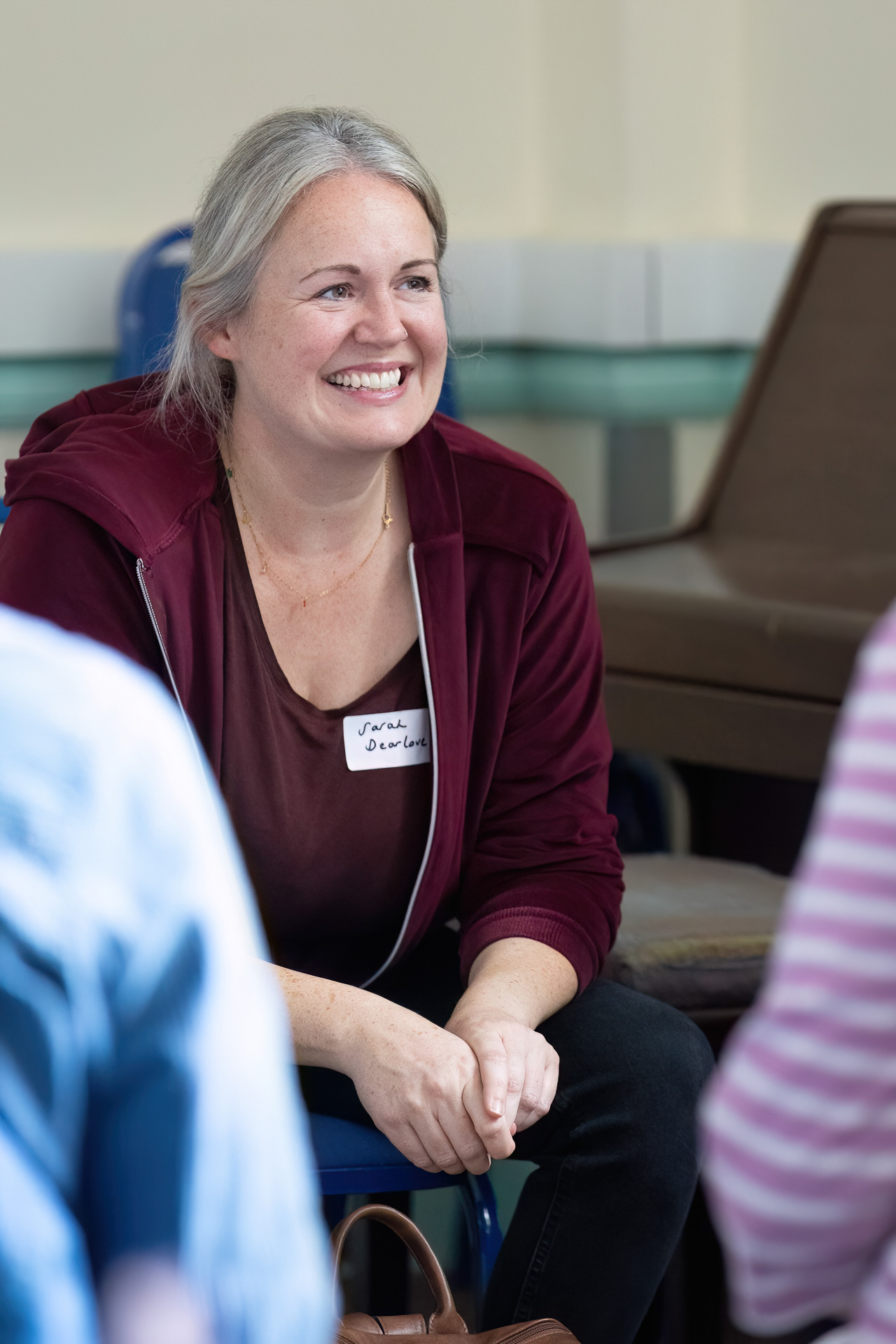 MMFTutorConference2026.83.web A smiling woman with grey hair sits indoors, wearing a maroon jacket and a name badge. She appears engaged in conversation with others in a casual, bright setting, embodying the welcoming spirit of the new TEMPO Ambassadors.