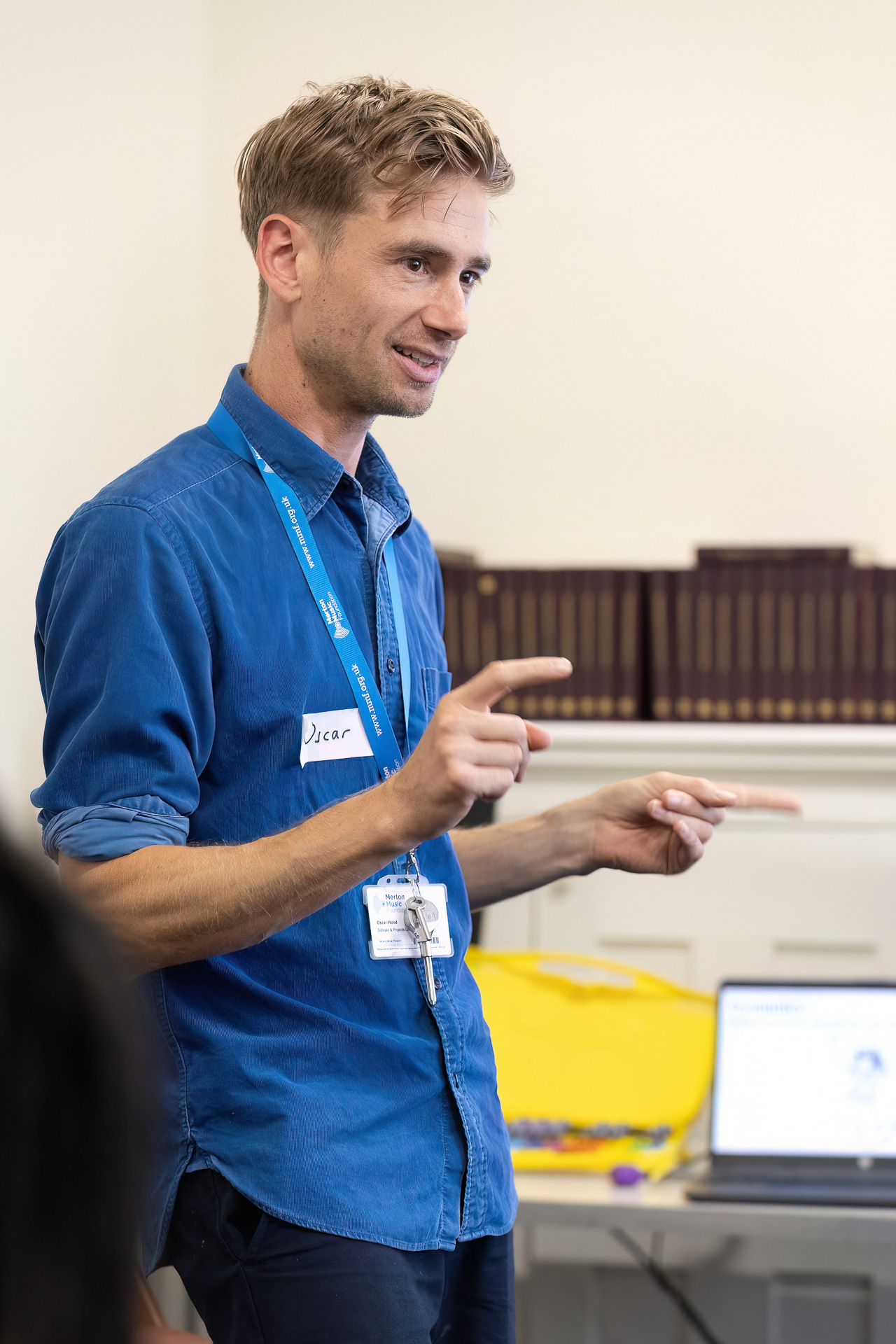 MMFTutorConference2026.85.web A man wearing a blue shirt and lanyard, one of the new TEMPO Ambassadors, gestures with his hands whilst speaking in a classroom setting. There are books on a shelf and an open laptop in the background.