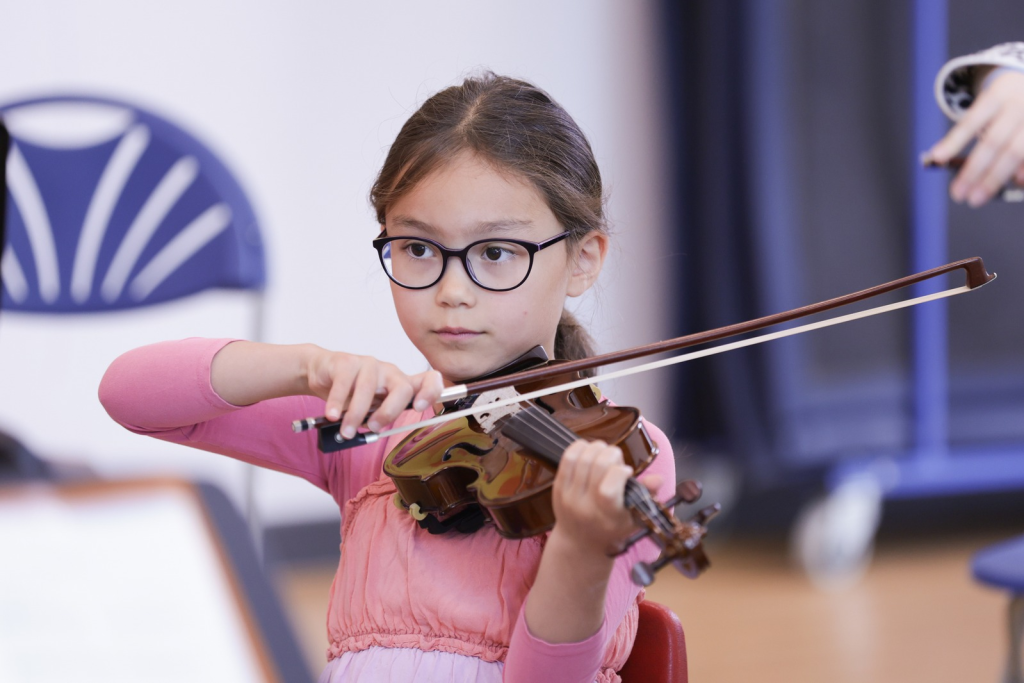 A young girl with glasses and brown hair, wearing a pink dress, plays the violin with concentration during her instrumental lessons. She is indoors, with a blue chair and blurred background.
