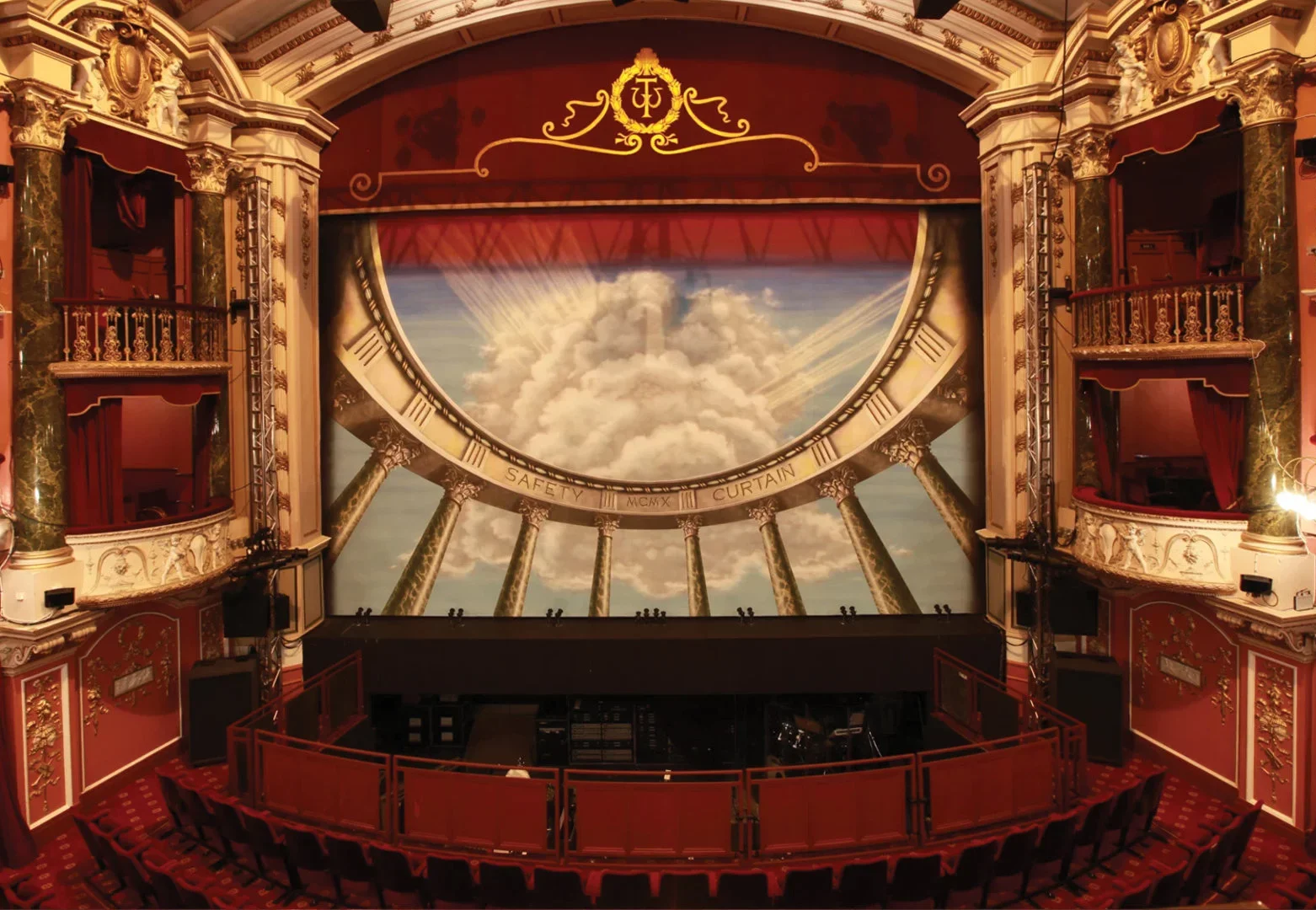 An ornate theatre with red seats and gold accents faces a stage with a painted backdrop showing clouds and sunlight, framed by two large columns and decorative balconies.