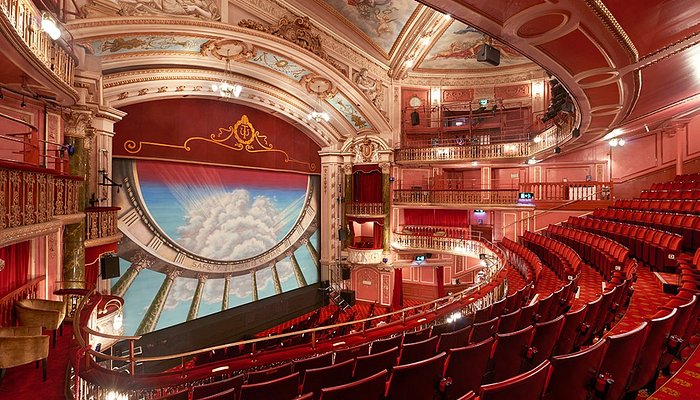Elegant, ornate theatre interior with red seats, gold accents, and detailed ceiling. The stage features a painted backdrop of clouds and sunlight. Multiple balconies overlook the main seating area.